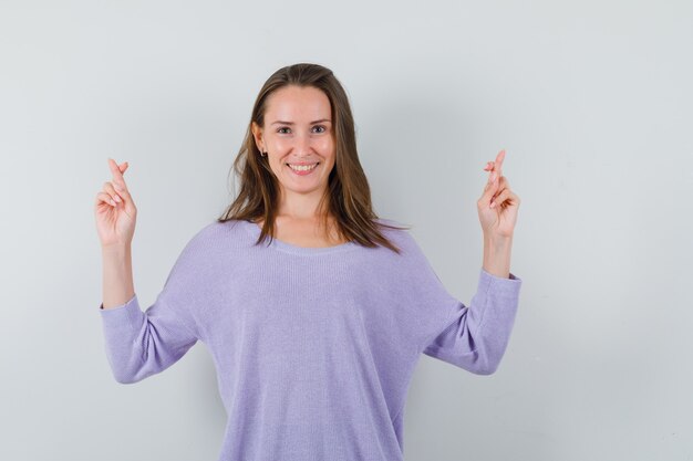 Young female keeping fingers crossed in lilac blouse and looking glad 