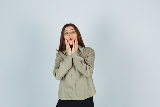 Young female keeping hands near open mouth in shirt, skirt and looking wondered. front view.