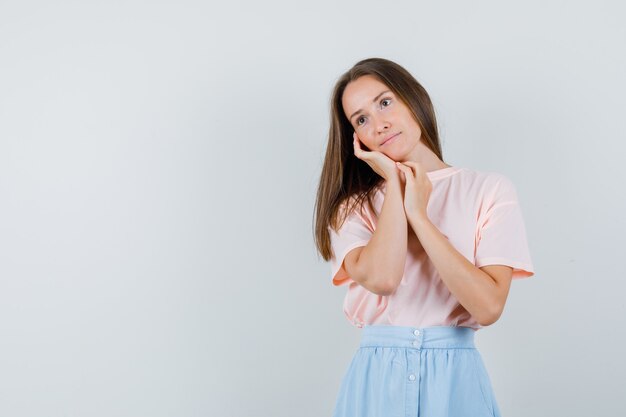 Young female leaning cheek on palm in t-shirt, skirt and looking pensive , front view.