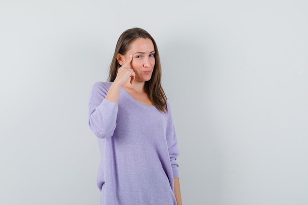 Young female in lilac blouse pointing at her temple and looking focused 