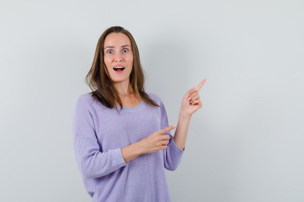 Young female in lilac blouse pointing at upper left corner while opening eyes widely and looking surprised 