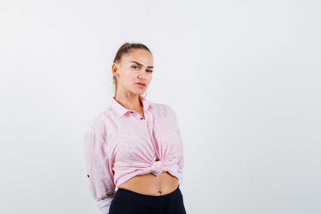 Young female looking at camera while holding hands behind back in casual shirt, pants and looking confident. front view.