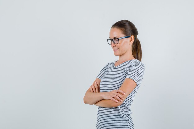 Young female looking down with crossed arms in t-shirt, glasses and looking pensive. front view.