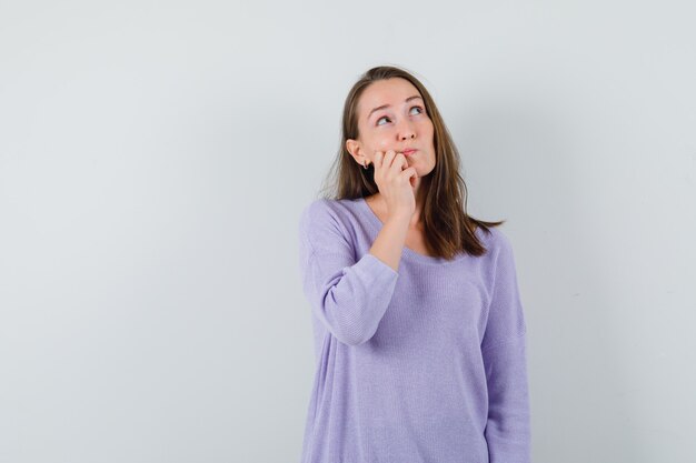 Young female looking upper left corner while thinking something in lilac blouse and looking puzzled 