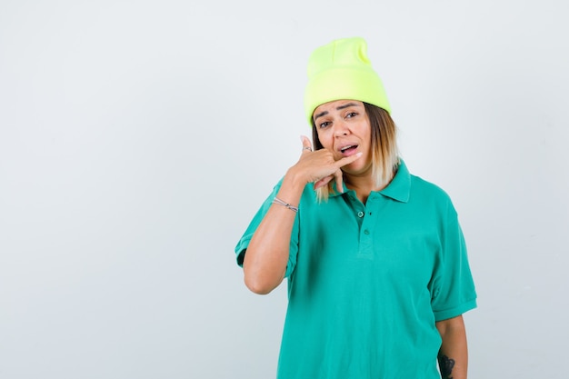 Young female making phone gesture in polo t-shirt, beanie and looking perplexed. front view.