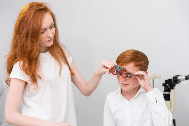 Free Photo young female optician wearing optometrist trial frame to cute boy for checking his eyes