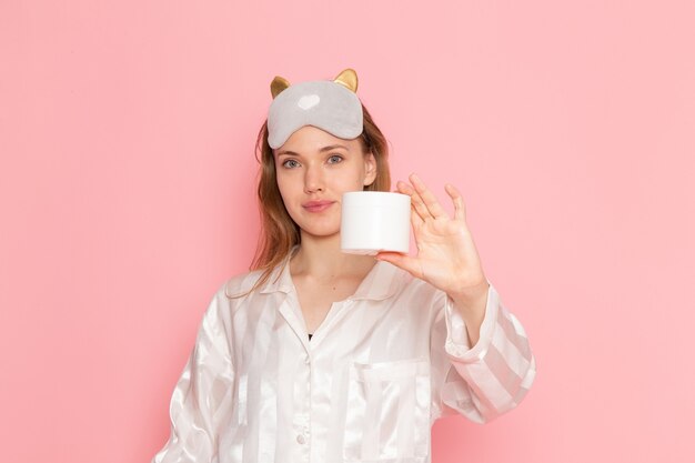 young female in pajamas and sleep mask holding cream with smile on pink