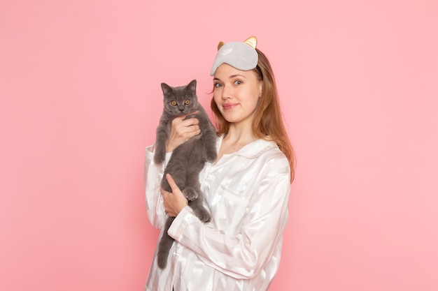 Free Photo young female in pajamas and sleep mask holding cute grey cat on pink