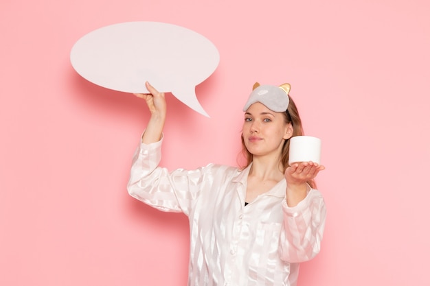 young female in pajamas and sleep mask holding white sign cream on pink