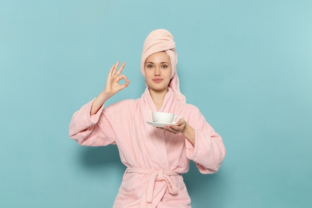 young female in pink bathrobe after shower holding cup of coffee on blue