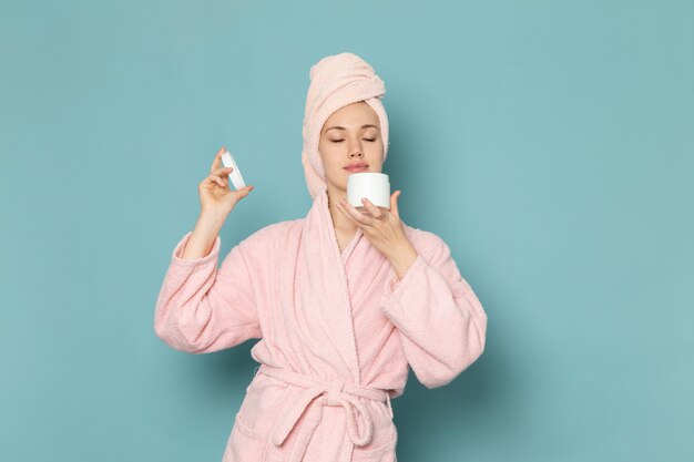 young female in pink bathrobe after shower holding and using cream on blue