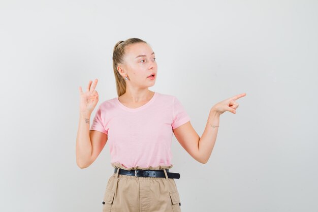 Young female pointing aside, showing ok sign in t-shirt, pants and looking focused. front view.