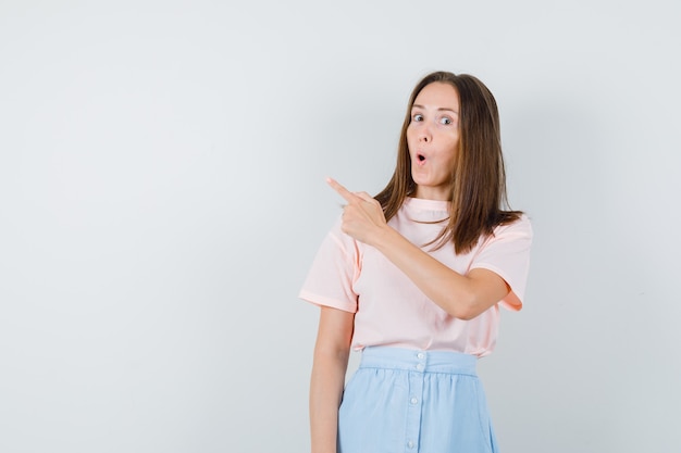 Young female pointing away in t-shirt, skirt and looking surprised , front view.