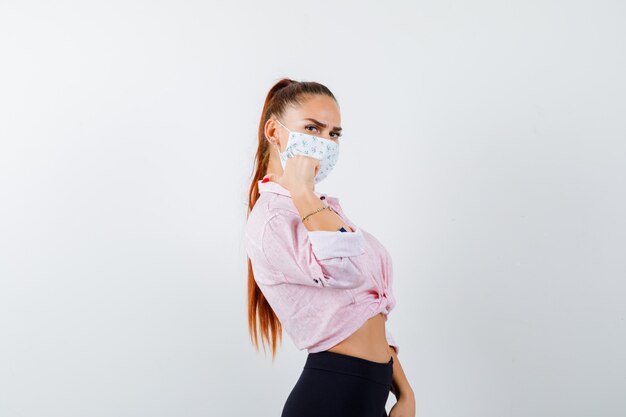 Young female pointing back with thumb in shirt, pants, mask and looking confident .