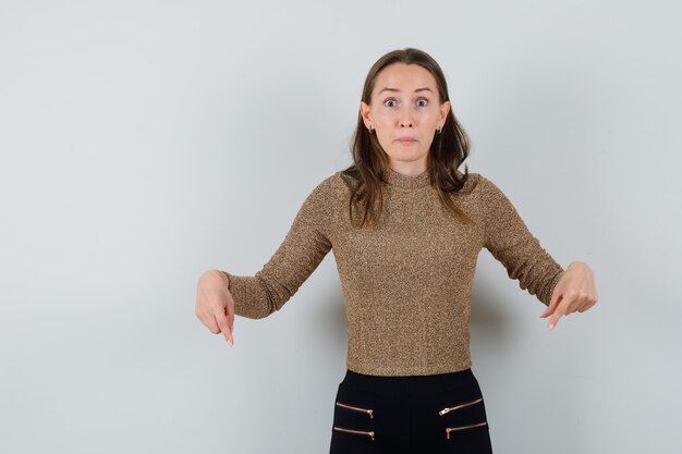 Young female pointing down in golden blouse and looking surprised , front view.