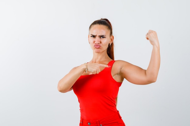 Young female pointing at muscles of arm in red tank top, pants and looking confident , front view.