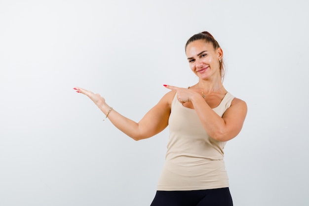 Young female pointing at palm in beige tank top and looking confident. front view.