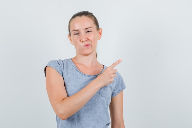 Young female pointing to side in grey t-shirt and looking displeased. front view.