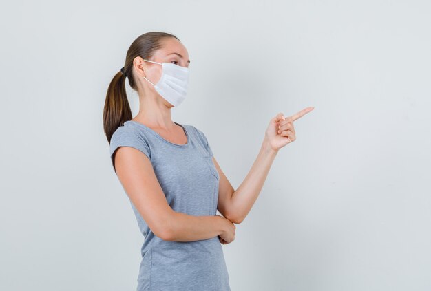 Young female pointing to side in grey t-shirt, mask and looking focused. front view.