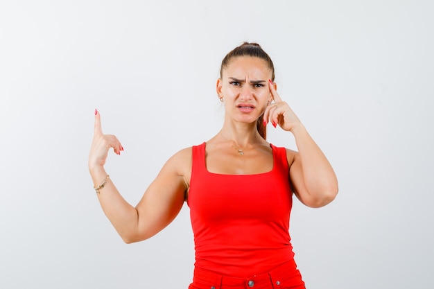 Young female pointing up in red tank top, pants and looking thoughtful. front view.