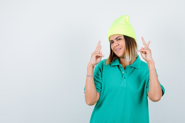 Young female in polo t-shirt, beanie showing victory gesture and looking cheerful , front view.