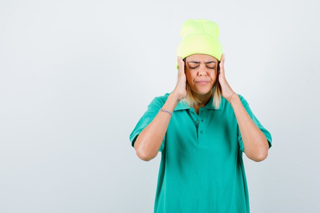 Young female in polo t-shirt, beanie with hands near face, closing eyes and looking focused , front view.