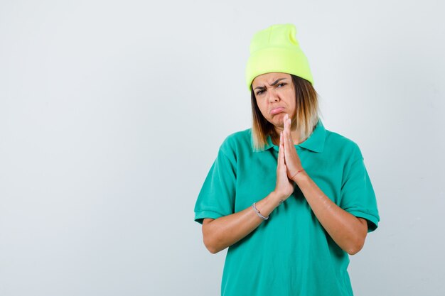 Young female in polo t-shirt, beanie with hands in praying gesture and looking upset , front view.