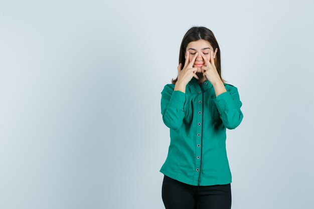 Young female pretending to rub face mask on around nose zone in green shirt, pants and looking calm. front view.