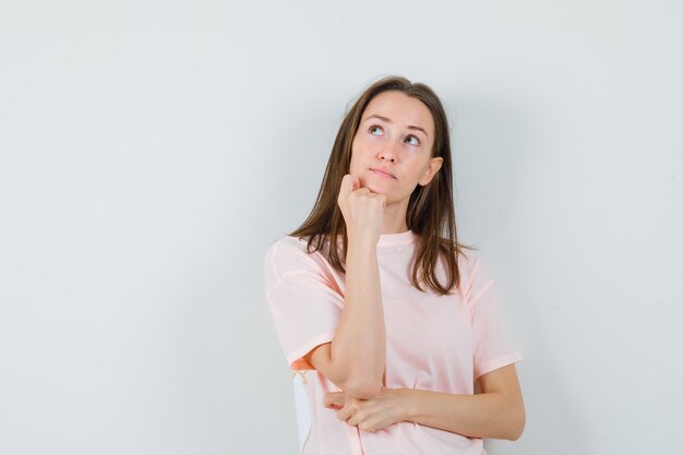 Young female propping chin on fist in pink t-shirt and looking pensive , front view.