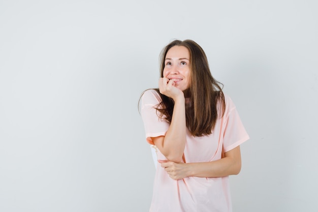 Young female propping chin on palm in pink t-shirt and looking dreamy. front view.