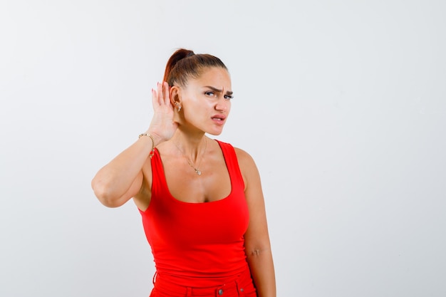 Young female in red tank top, pants holding hand behind ear and looking focused , front view.