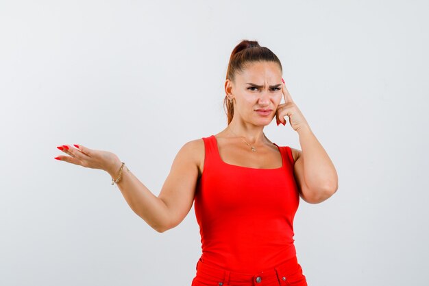 Young female in red tank top, pants stretching hand in questioning gesture and looking pensive , front view.