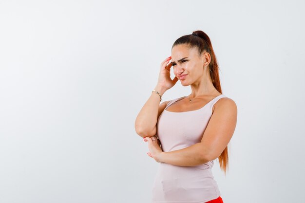 Young female rubbing temple in white tank top and looking exhausted , front view.