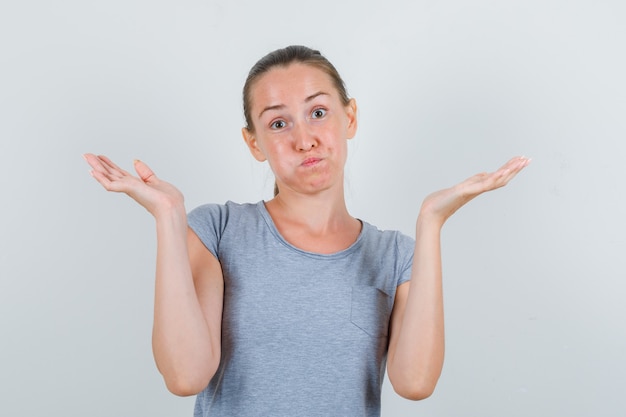 Young female showing helpless gesture in grey t-shirt and looking confused. front view.