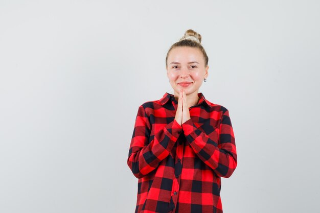 Young female showing namaste gesture in checked shirt and looking merry , front view.