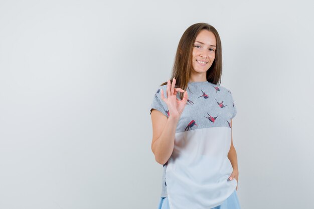 Young female showing ok gesture in t-shirt, skirt and looking cheerful , front view.