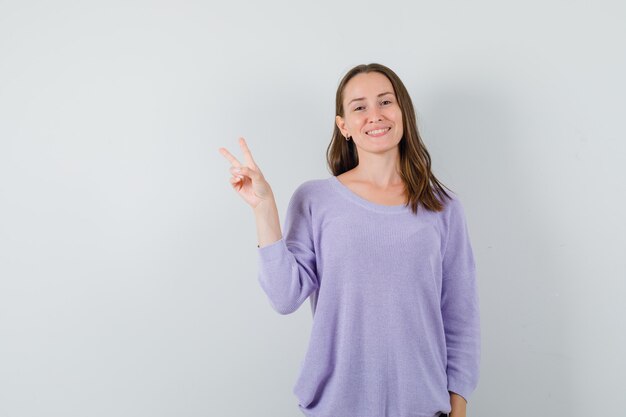 Young female showing V-sign in lilac blouse and looking joyful 