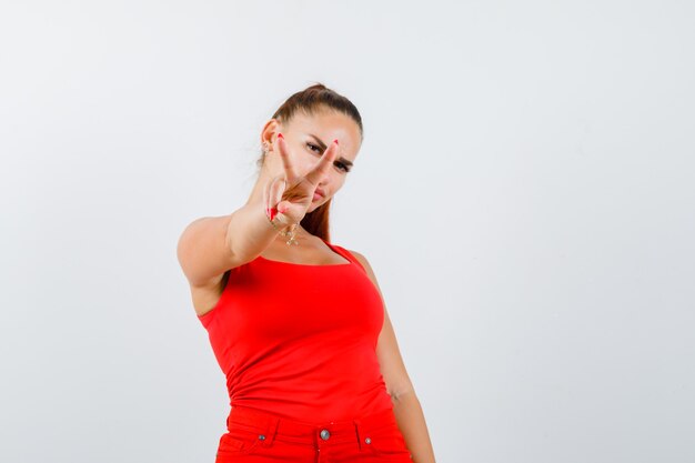 Young female showing victory sign in red tank top, pants and looking lucky , front view.