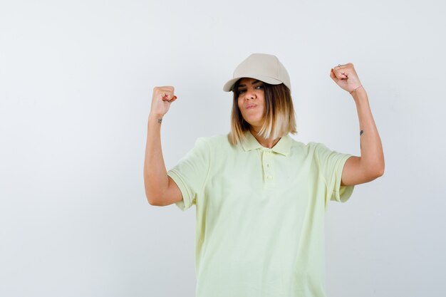 Young female showing winner gesture in t-shirt, cap and looking lucky , front view.