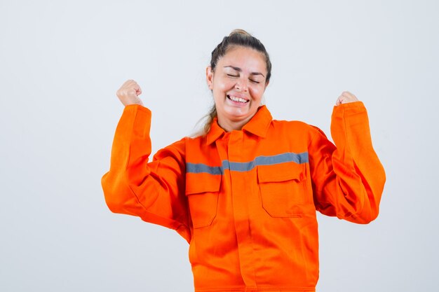 Young female showing winner gesture in worker uniform and looking cheerful , front view.