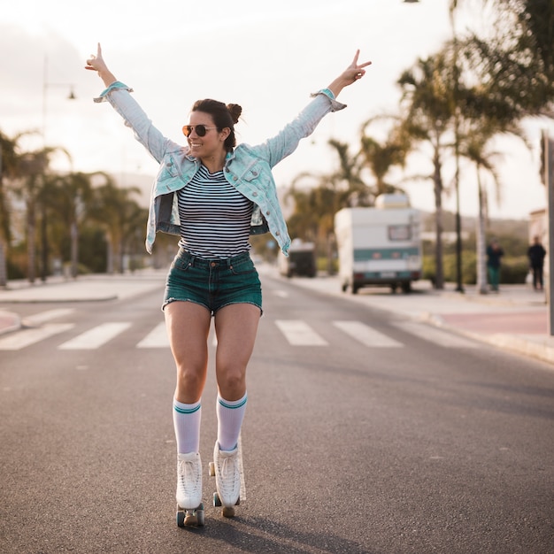 Free photo young female skater raising her arms making peace gesture balancing on road