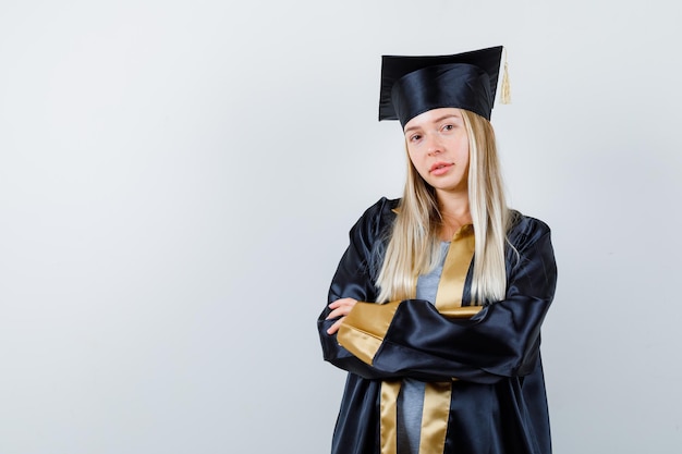 Young female standing with crossed arms in graduate uniform and looking confident