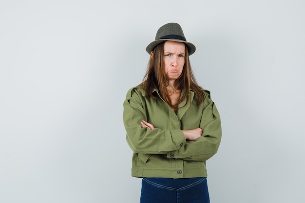 Free photo young female standing with crossed arms in jacket, pants, hat and looking offended , front view.