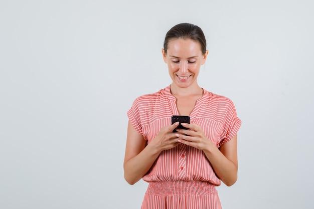 Young female in striped dress typing on mobile phone and looking glad , front view.