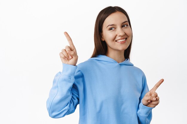 Young female student in blue hoodie, pointing sideways, two choices ways, showing variants of items on sale, smiling and lookin left at one side, standing over white wall.