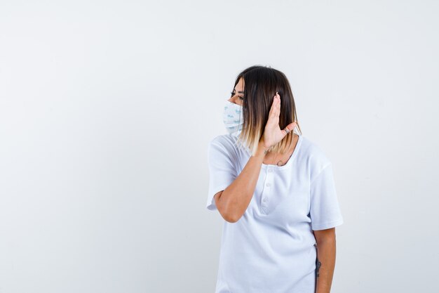 Young female in t-shirt, mask holding hand behind ear and looking wondered , front view.