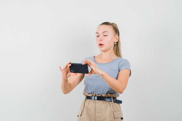 Young female taking photo on mobile phone in t-shirt, pants , front view.