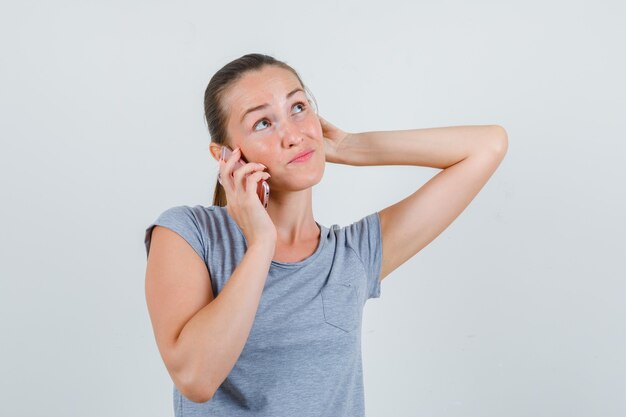 Young female talking on mobile phone in grey t-shirt and looking hesitant , front view.