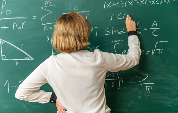 Free Photo young female teacher standing in front blackboard writing something on blackboard with stranded for board in classroom