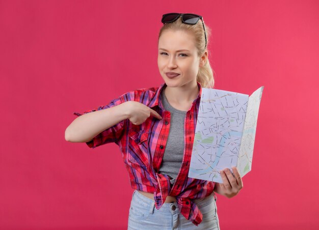 Young female traveler wearing red shirt and glasses on her head holding map points to herself on isolated pink wall